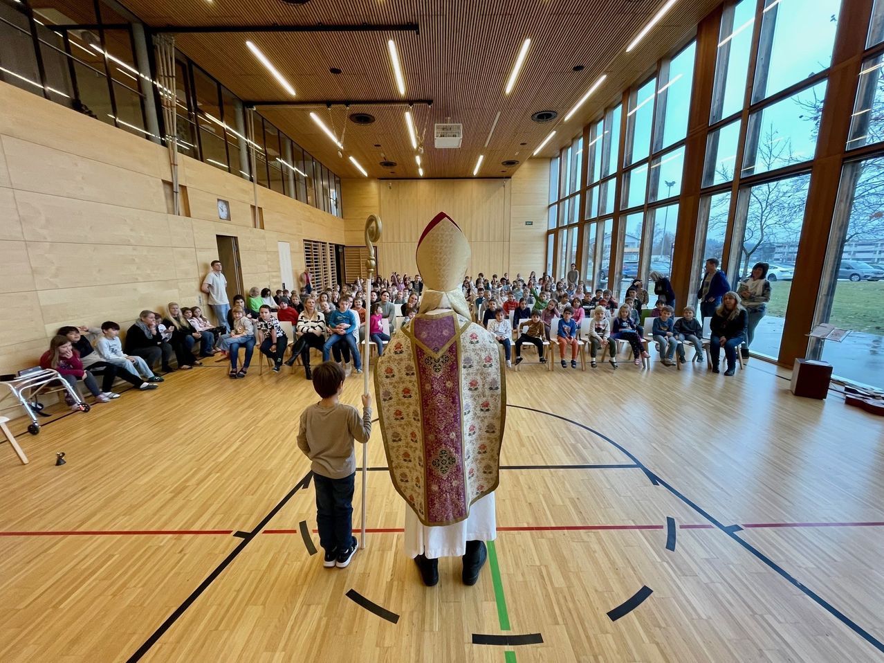Ein Priester steht in einer Turnhalle mit einem kleinen Jungen, umgeben von einem Publikum sitzender Kinder. Der Raum hat Holzböden und große Fenster.