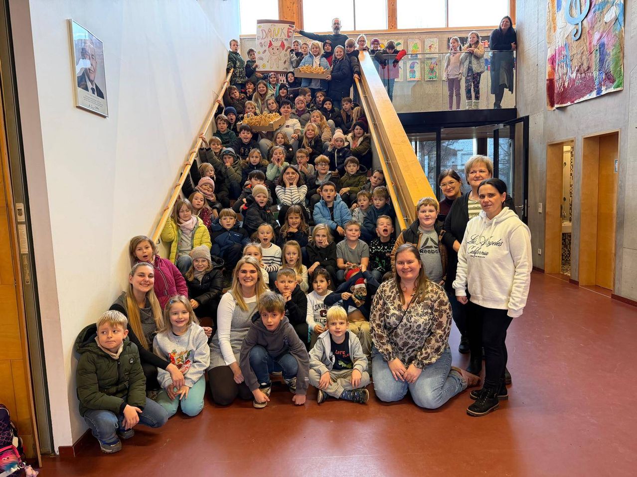 Eine Gruppe von Kindern und Erwachsenen versammelt sich auf einer Treppe, einige sitzen und andere stehen. Eine Person hält ein Banner, während andere Tabletts mit Essen halten. Erwachsene stehen am Fuße der Treppe.