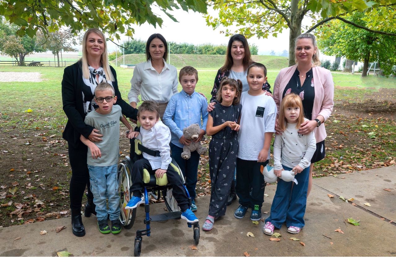 Eine Gruppe von Menschen, darunter Erwachsene und Kinder, steht in einem Park. Einige Kinder halten Kuscheltiere. Ein Kind im Rollstuhl schaut nach links.