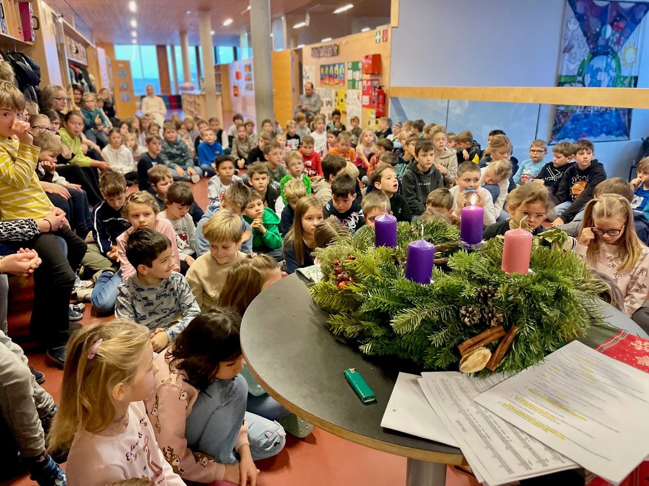 Ein Klassenzimmer voller Kinder, die auf dem Boden sitzen und einer Veranstaltung zuschauen. Ein Tisch davor hat eine Adventskrone mit Kerzen und einigen Papieren.