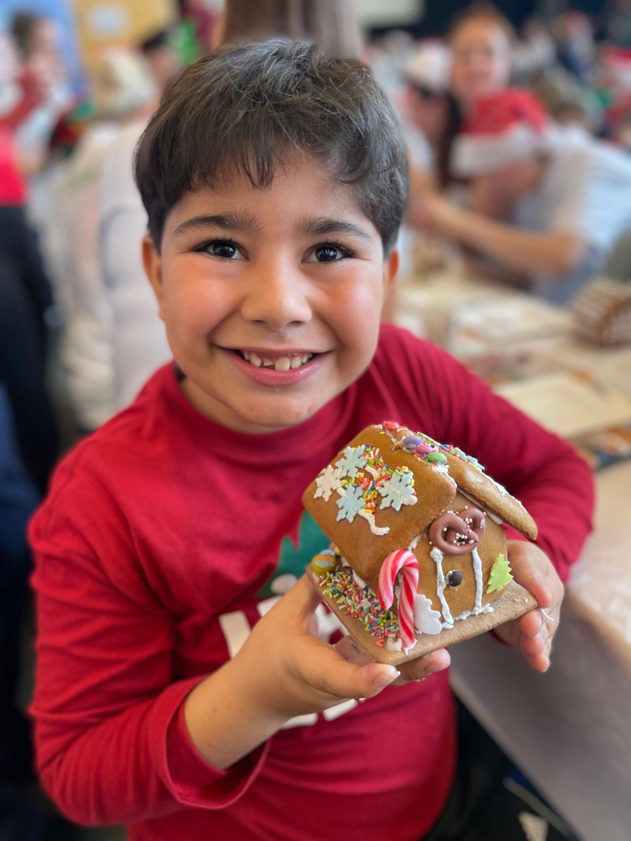 Ein junger Junge in einem roten Shirt lächelt und hält ein Lebkuchenhaus mit Süßigkeiten und Zuckerguss Dekorationen.