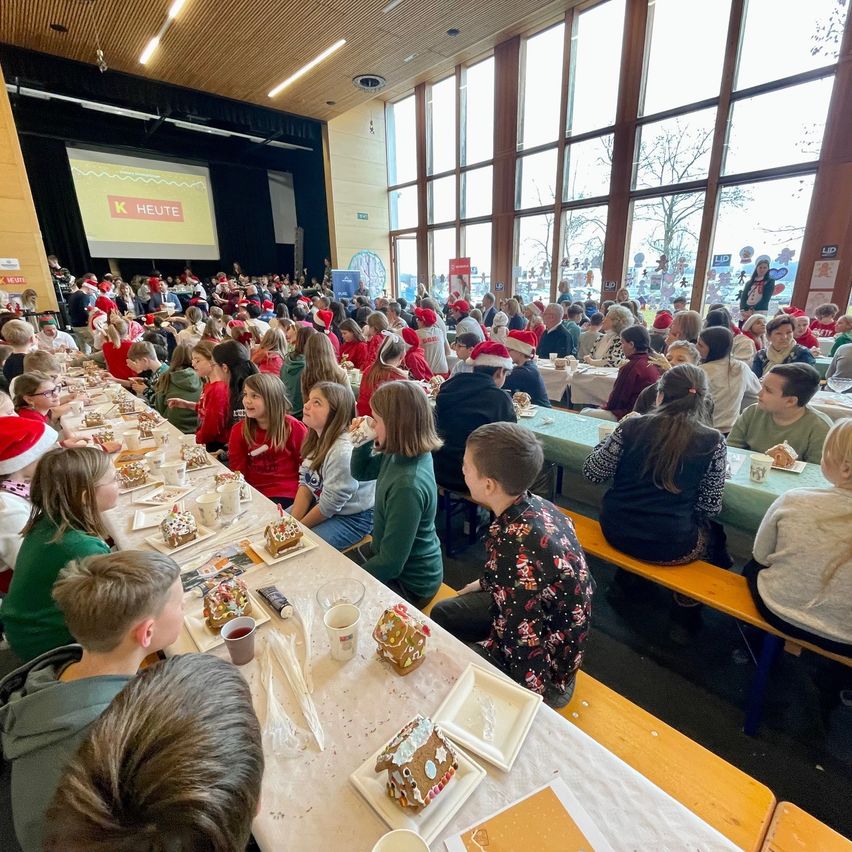 Eine große Gruppe von Menschen, wahrscheinlich Schüler, sitzen an langen Tischen in einem Raum, möglicherweise einer Schulmensa, essen und plaudern. Der Raum hat große Fenster und eine Projektionsleinwand zeigt etwas im Hintergrund. Einige Schüler tragen Weihnachtsmützen.