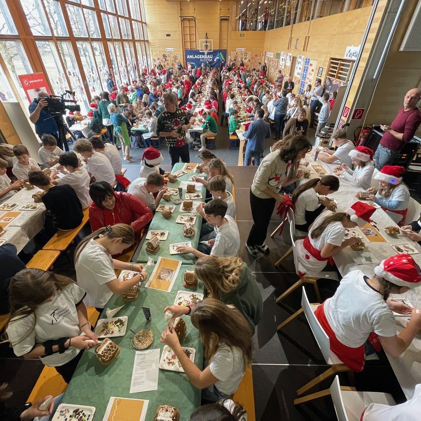 Eine Gruppe von Menschen sitzt um lange Tische in einem großen Raum herum, trägt Weihnachtsmützen und weiße Hemden. Sie essen von Tellern. Einige sind Erwachsene, andere Kinder. Es gibt gedeckte Tische mit Dekorationen, und ein Kameramann filmt.