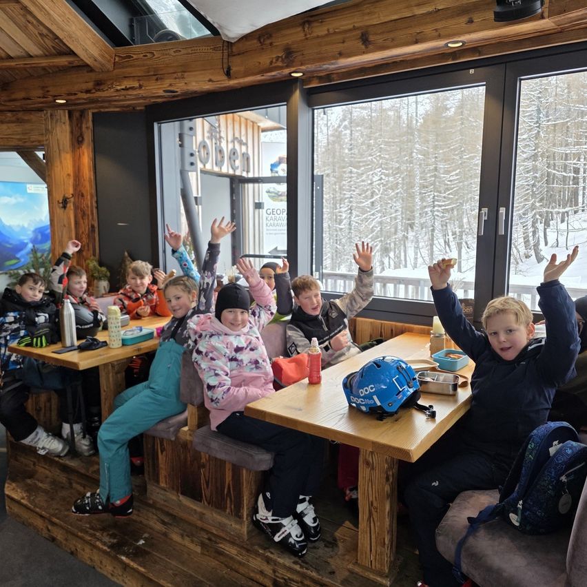 Mehrere Kinder, wahrscheinlich auf einer Skifahrt, sitzen an Tischen in einer gemütlichen, hölzernen Lodge. Sie haben alle die Hände erhoben, und einige tragen Skiausrüstung. Die Lodge hat große Fenster, die eine verschneite Landschaft draußen zeigen.