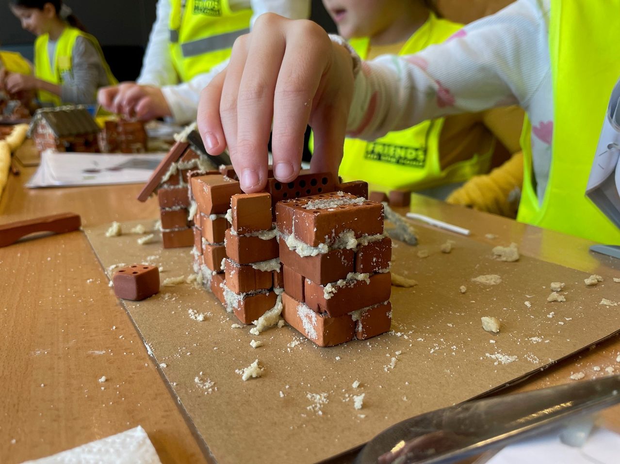 Ein Kind in einer gelben Sicherheitsweste platziert einen Ziegelstein auf einen Stapel von Ziegelsteinen auf einem Tisch.