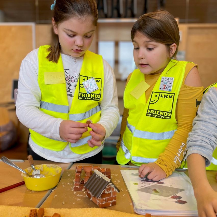 Zwei Mädchen in neonfarbenen Sicherheitswesten arbeiten an einem Miniatur-Ziegelhaus. Ein Mädchen verwendet einen Pinsel, um Mörtel aufzutragen, während das andere Steine hinzufügt. In der Nähe befindet sich eine gelbe Schüssel mit Mörtel. Sie sind von weiteren Steinen und einer Karte auf einem Tisch umgeben.