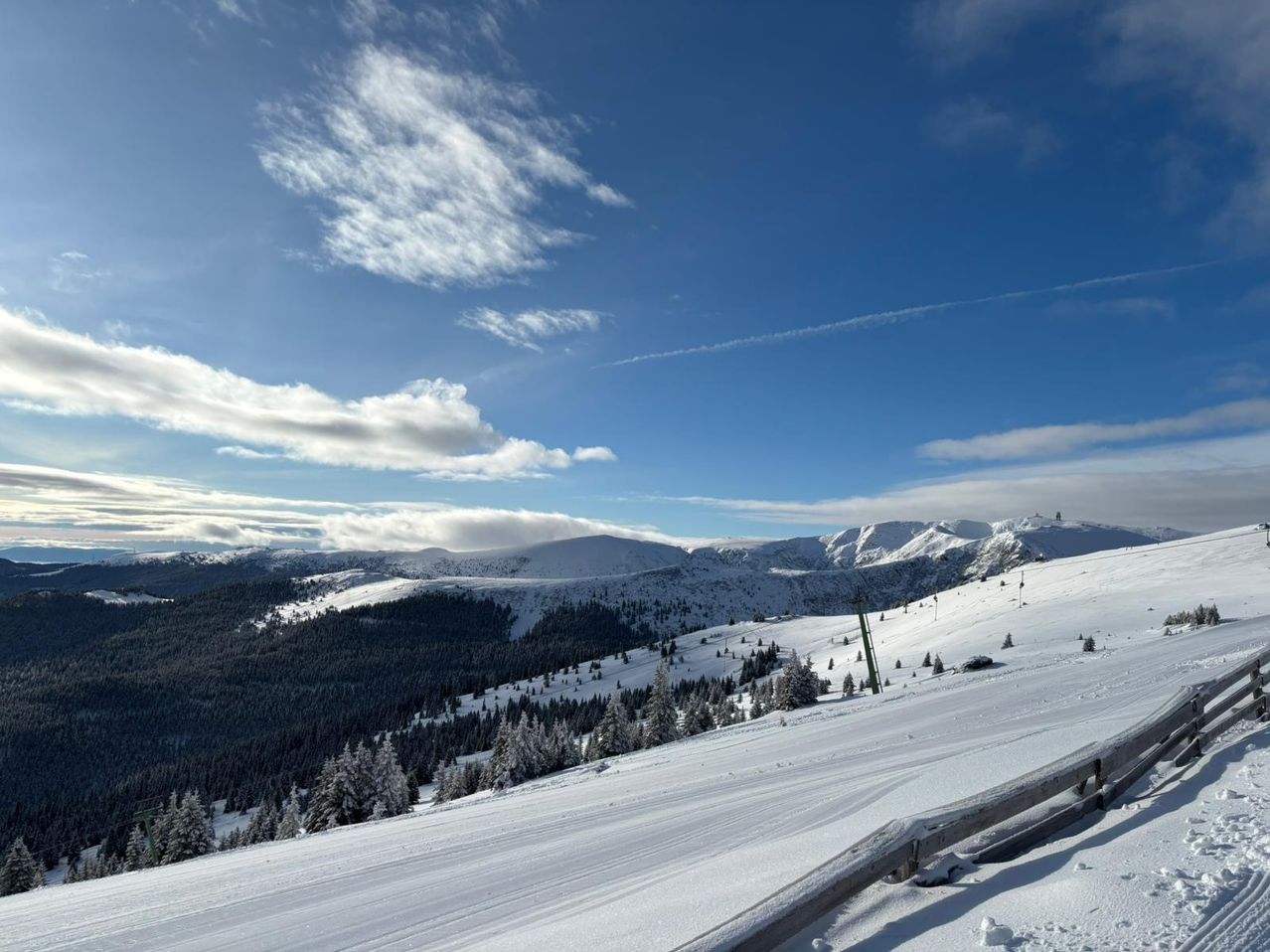 Eine verschneite Berglandschaft mit Kiefern, blauem Himmel und Wolken. Rechts ist ein Holzzaun.