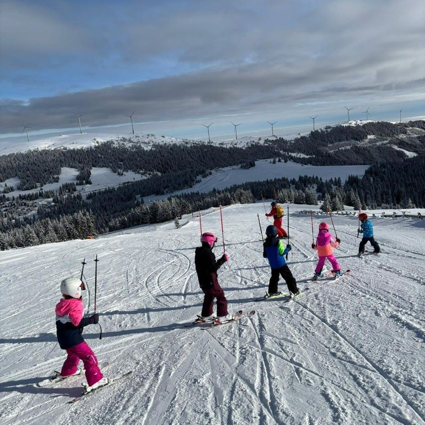 Mehrere Kinder fahren mit Skiern einen schneebedeckten Hang hinab, mit Windturbinen im Hintergrund.