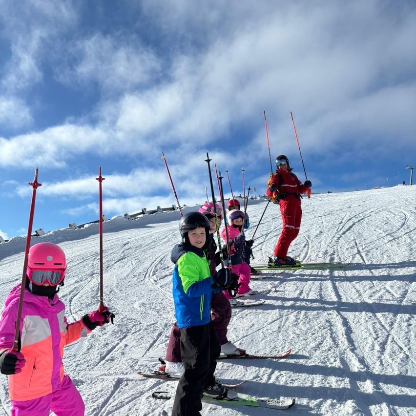 Eine Gruppe junger Skifahrer steht auf einem verschneiten Hang. Sie halten Skistöcke und tragen Helme und Skianzüge. Ein Erwachsener in roter Skiausrüstung steht hinter ihnen.