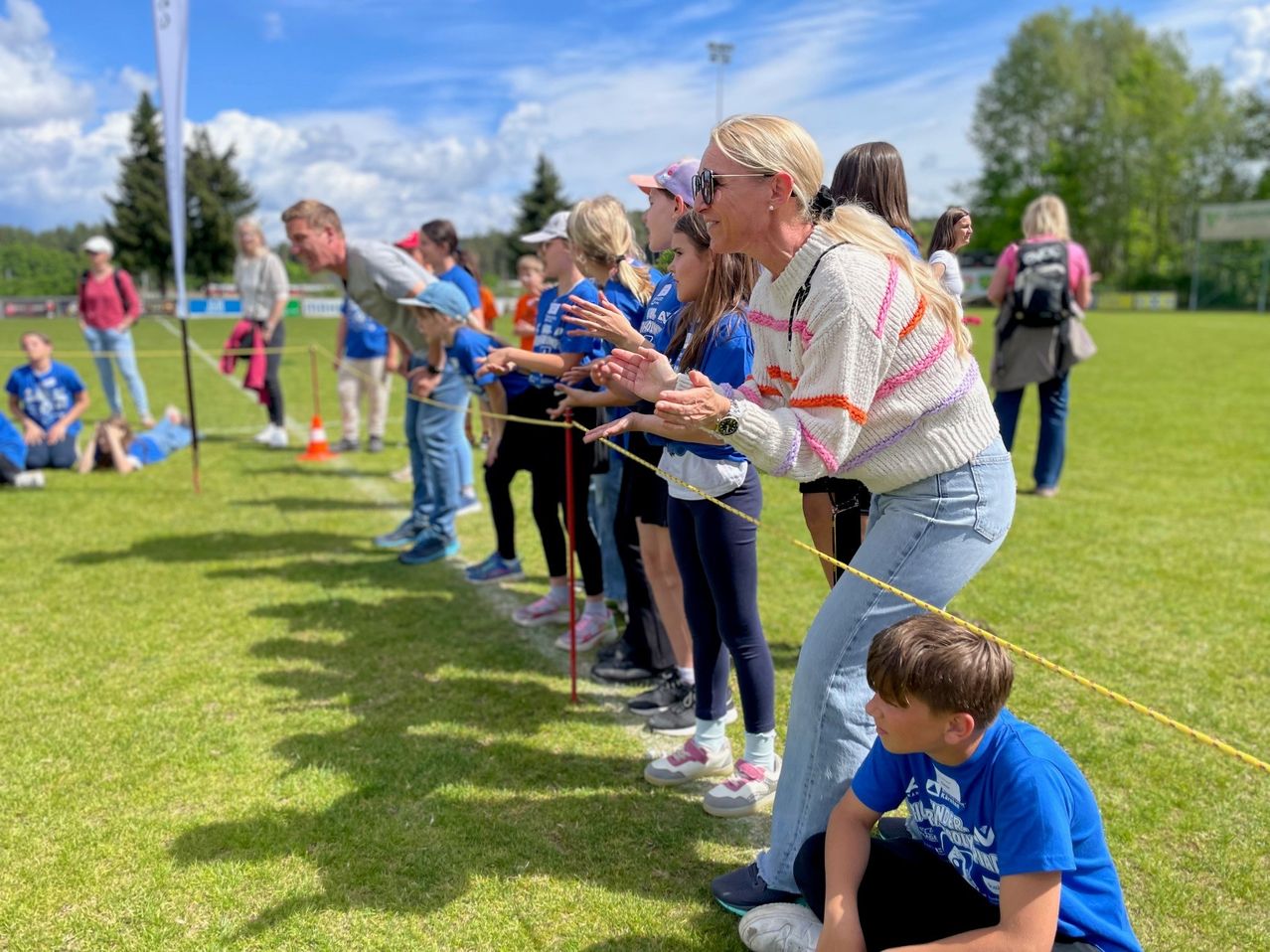 A group of people in blue shirts stand in line on a grassy field. An adult woman in a white sweater and jeans speaks to them. Nearby, a boy sits on the ground.