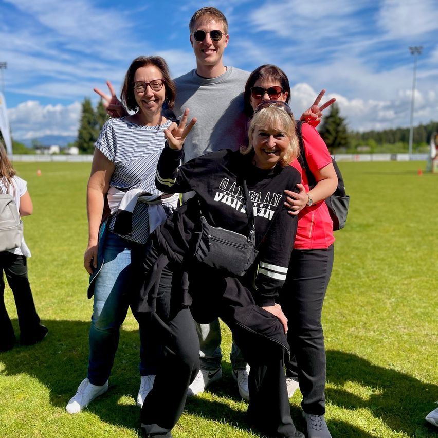 Five people pose for a picture on a grassy field. The man and woman in the center make peace signs. Behind them are trees, a streetlight, and a blue sky with clouds.