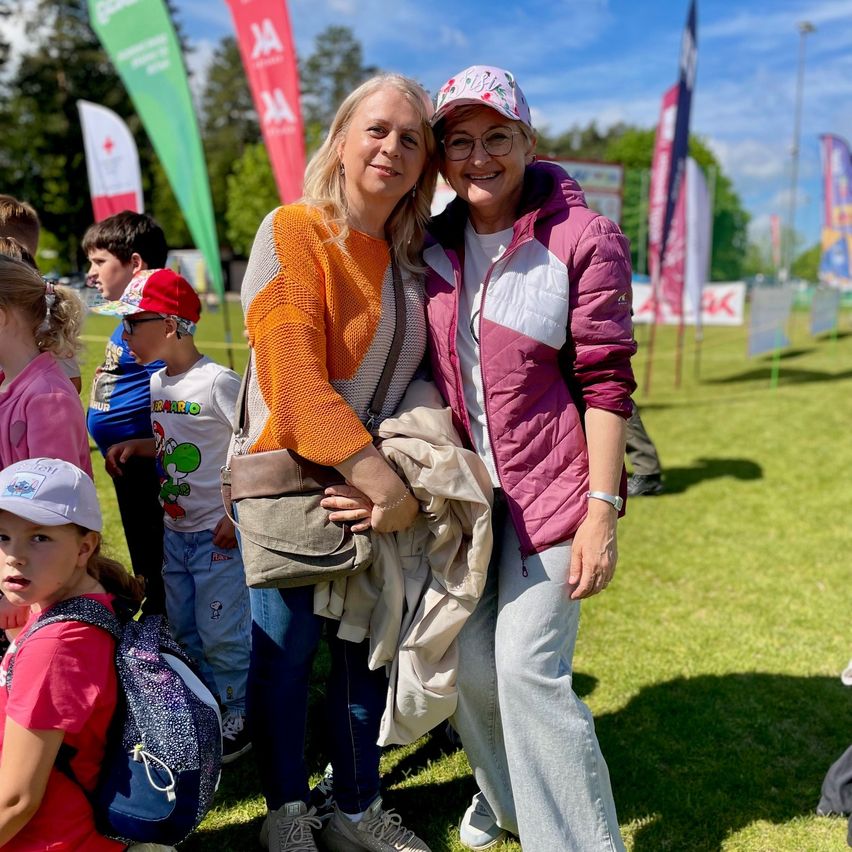 Two women smiling in a park with children and flags in the background. One wears a hat and glasses, the other a necklace and glasses.