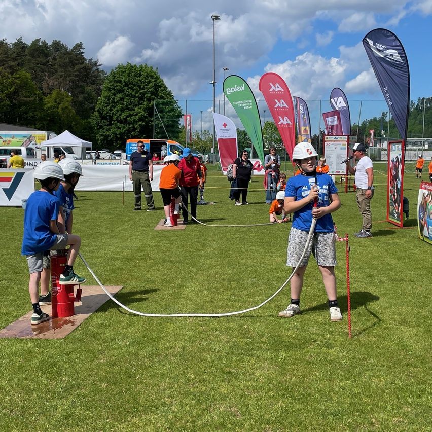 A group of children wearing helmets are participating in a fire extinguisher training on a grassy field with banners and tents in the background.