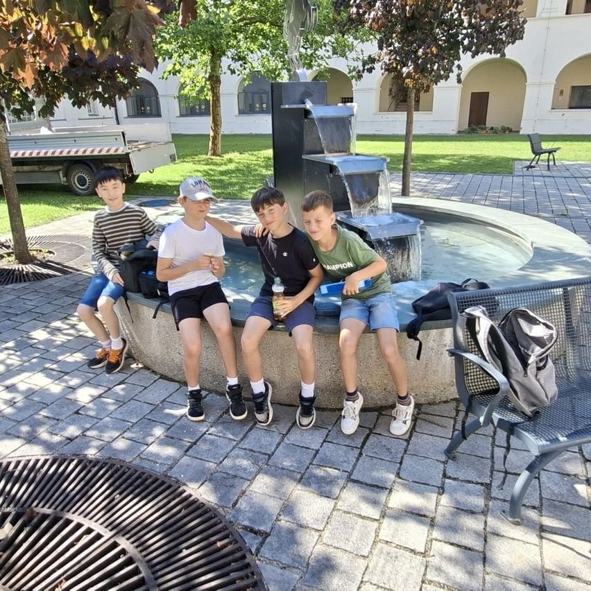 Vier Jungen sitzen um einen Brunnen in einem Park. Einer hält eine Flasche, ein anderer einen blauen Behälter. Bäume und ein Gebäude sind im Hintergrund.