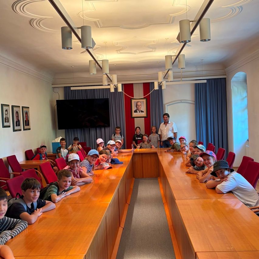 Eine Gruppe von Kindern sitzt mit Erwachsenen in einem Konferenzraum. Ein großer Fernseher ist an der Wand, und darüber befindet sich ein Bilderrahmen.