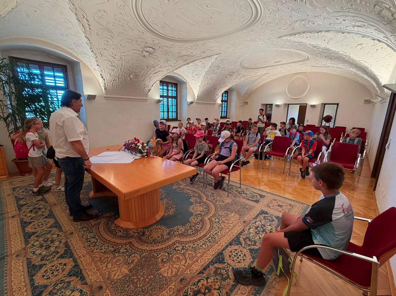 Eine Gruppe von Schülern sitzt in roten Stühlen in einem großen Saal mit kunstvollen Deckendesigns. Ein Mann steht an einem Tisch mit einer Blumenarrangement.