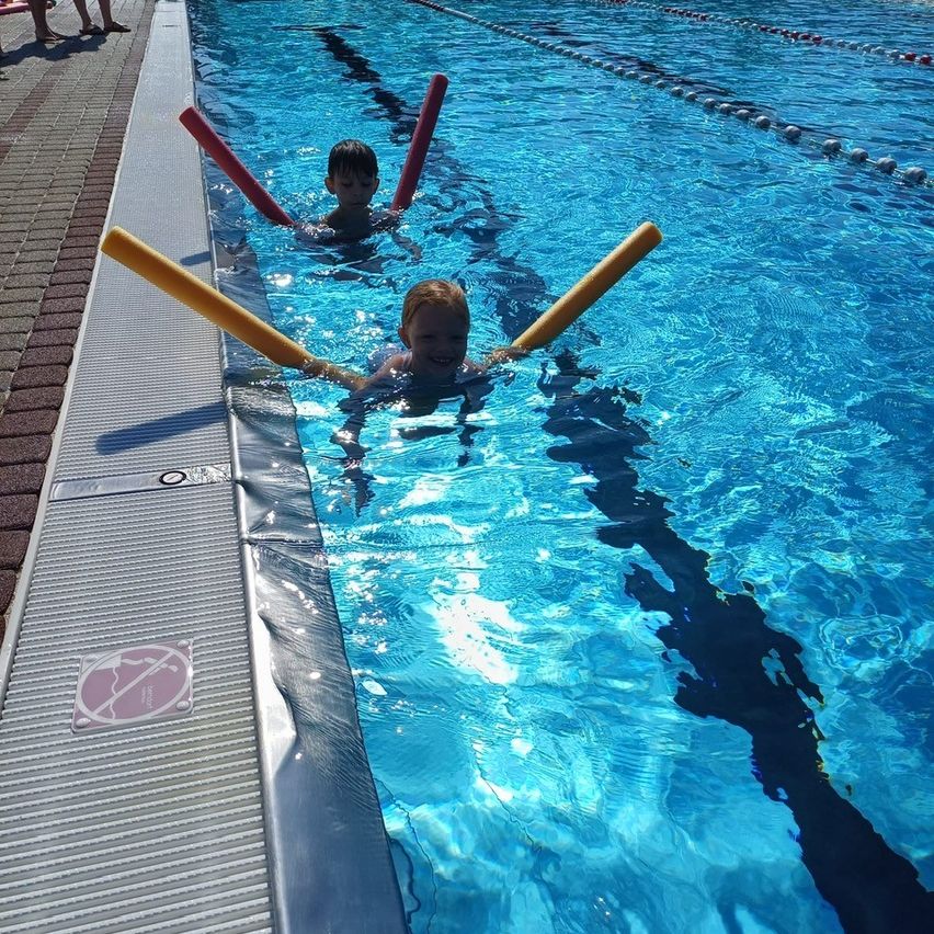 Zwei kleine Kinder schwimmen in einem Pool mit Schwimmhilfen. Der Pool hat Bahnen und ist von einem Ziegelrand umgeben.