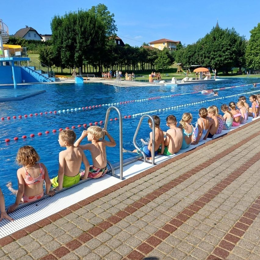 Kinder und Jugendliche warten am Rand eines Schwimmbeckens in einer Außenpoolanlage. Bäume und Häuser sind im Hintergrund sichtbar.