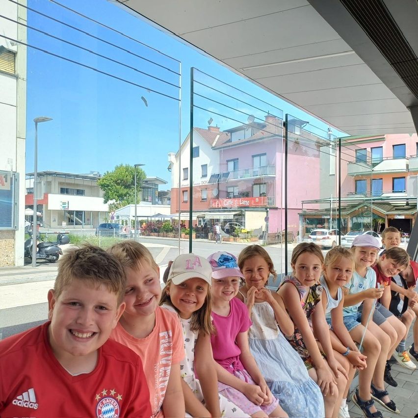 Eine Gruppe lächelnder Kinder in Hüten posiert für ein Foto auf einer Bank unter einer überdachten Überführung. Dahinter sind Gebäude und Autos zu sehen.