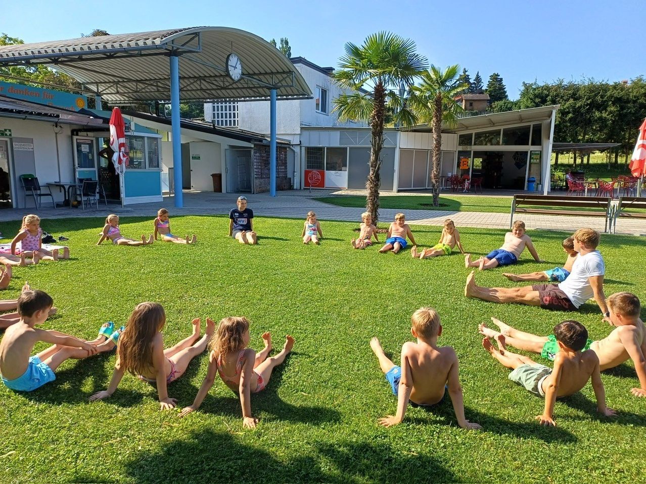 Kinder sitzen in einem Kreis auf einem Rasen, möglicherweise in der Pause. Hinter ihnen steht ein Gebäude mit Glastüren und Fenstern. Es gibt Bäume und einen Mülleimer in der Umgebung.