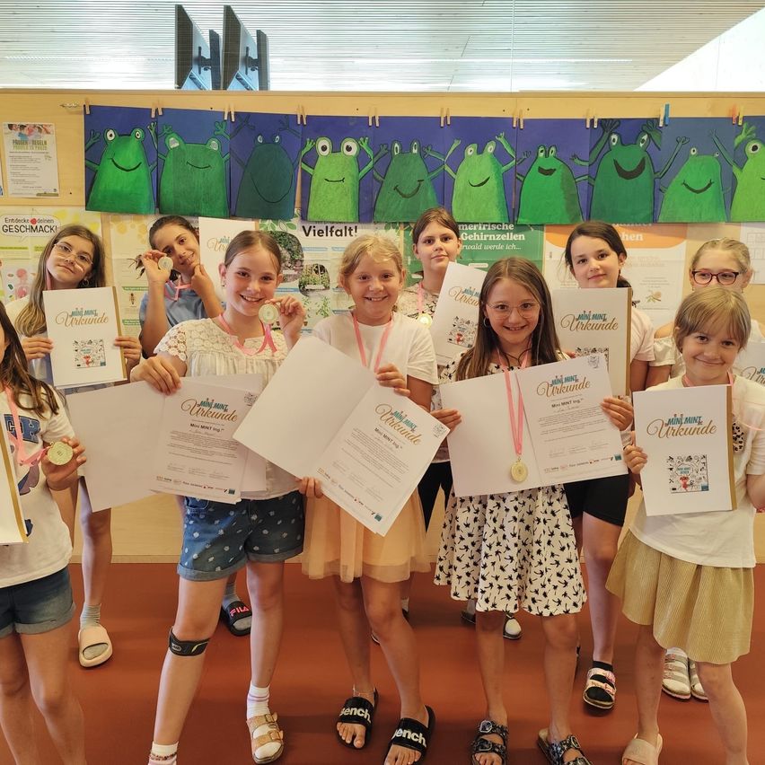 Eine Gruppe junger Mädchen in einem Klassenzimmer hält Zertifikate, Medaillen und Bücher in der Hand. Sie lächeln und stehen in einer Reihe. Hinter ihnen sind Poster mit Froschzeichnungen an der Wand befestigt.