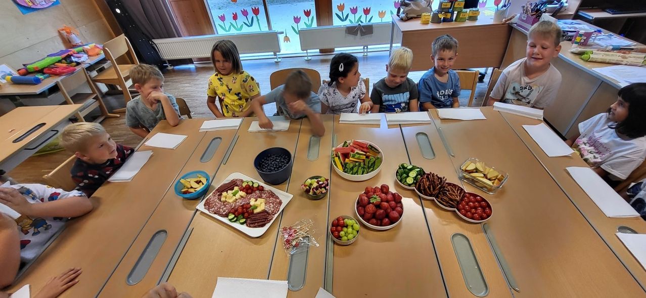 Several children sit at a table with various foods. Bowls of fruits, vegetables, and meats are on the table. The children look at the camera.