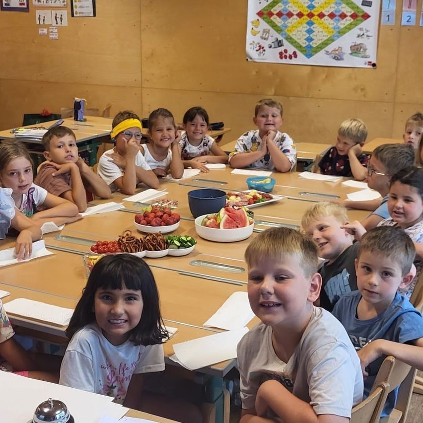 A classroom filled with young children around a table with food, smiling and posing for a photo. There are posters on the wall.