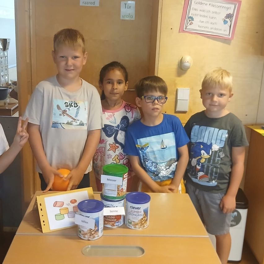 A group of children are standing around a table with various food items and labels, possibly in a classroom.