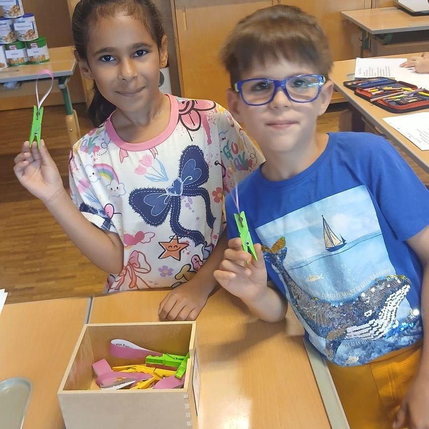Two kids are sitting at a desk. A girl holds a green paper clip and a boy holds a green paper clip with a sailboat on his shirt.