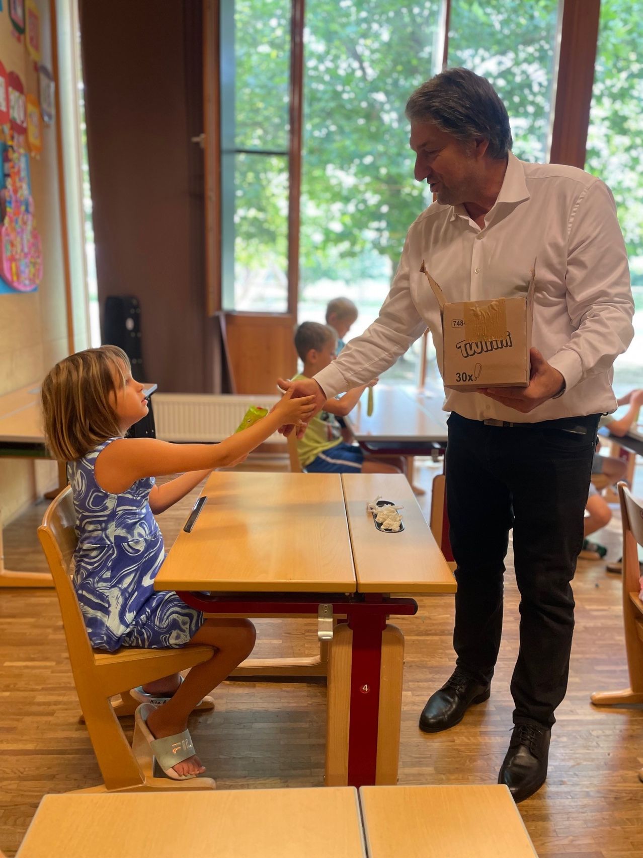 A man in a white shirt hands a box to a young girl sitting at a desk in a classroom.