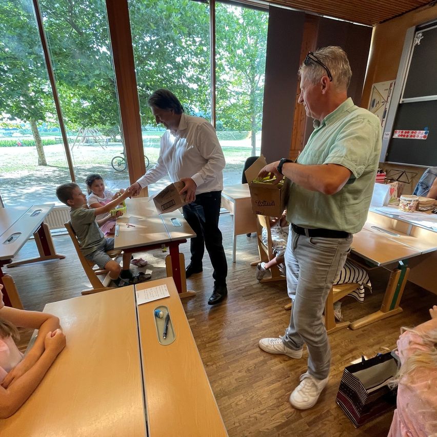 Two men in a classroom are giving boxes to children sitting at desks. The children are smiling and holding the boxes.