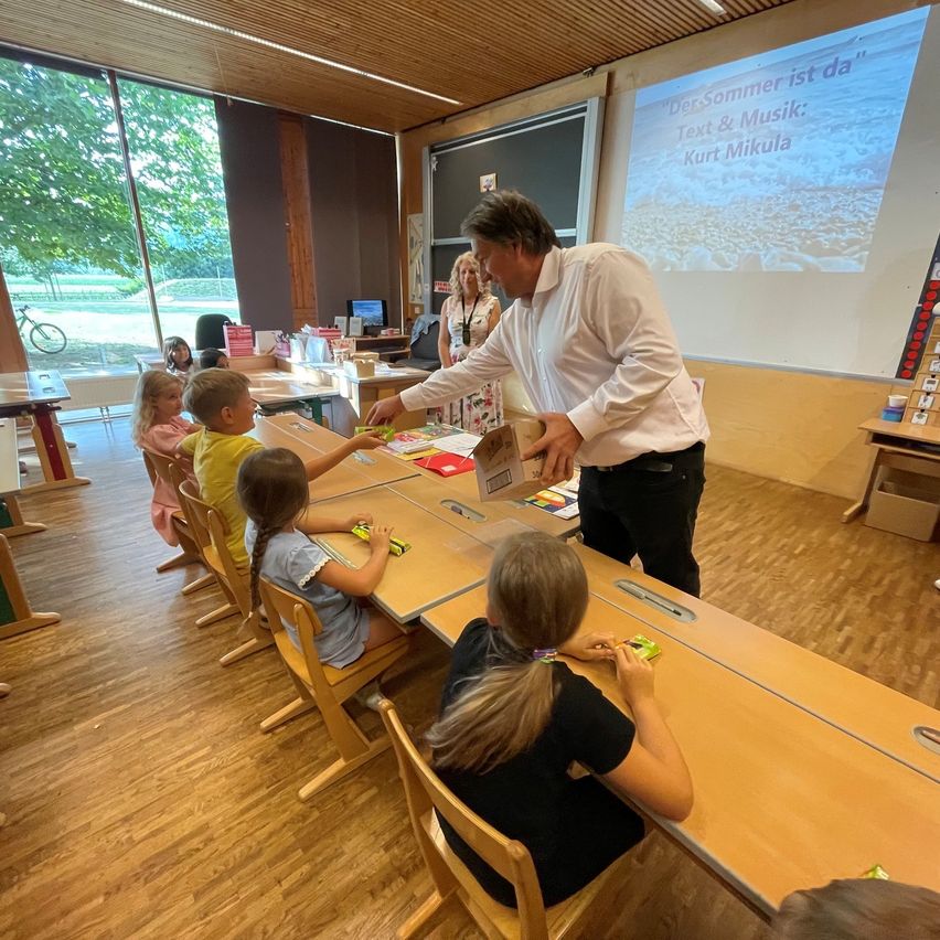 A man in a white shirt is teaching a class of young children seated at a long table, with a projector screen in the background displaying a presentation.