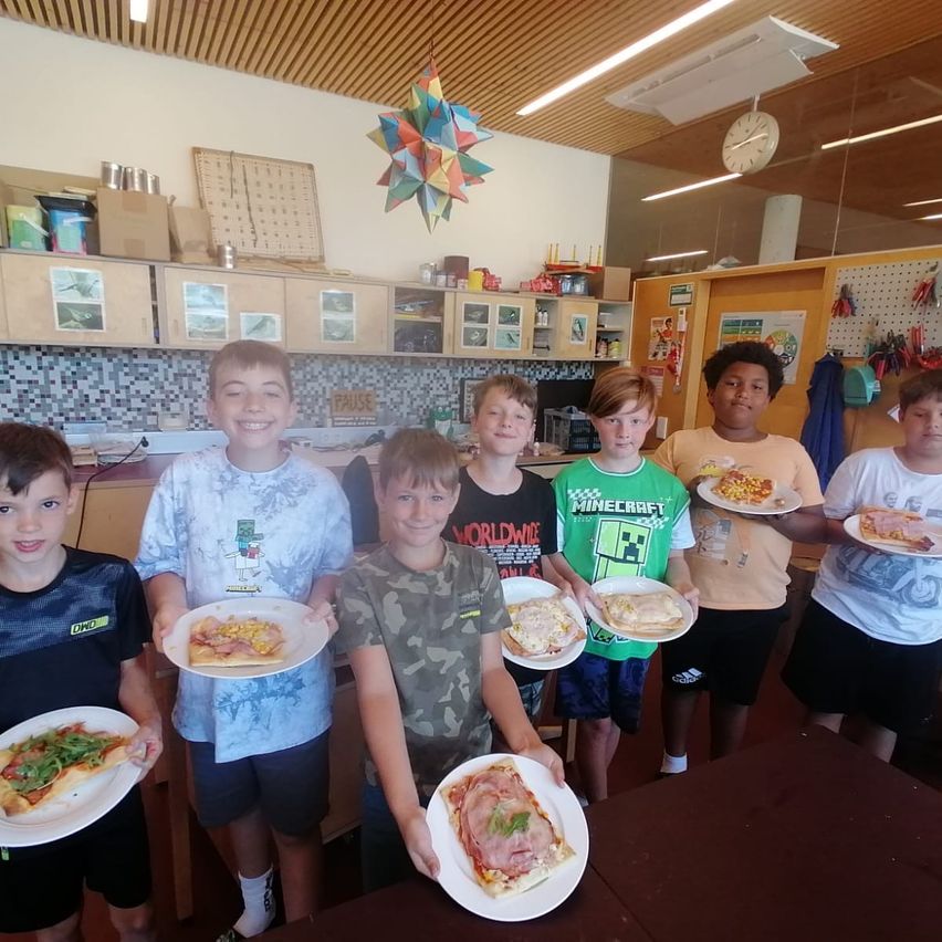 A group of young boys in a classroom setting, each holding a plate of pizza. They are smiling and posing for a photo.