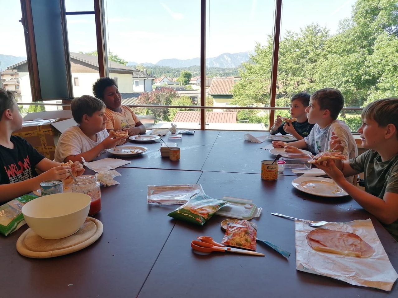 A group of children sitting around a table eating pizza, with a view of houses and mountains outside the window.
