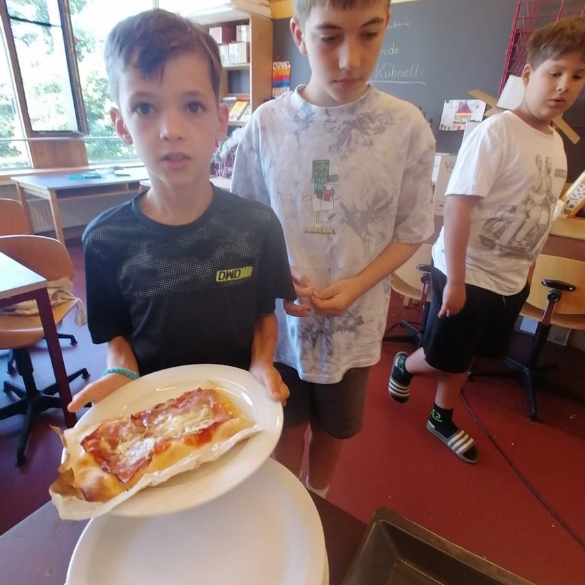 Three boys stand in a classroom, one holding a plate with a slice of pizza. The boy in the middle wears a white shirt with a graphic, while the others wear black shirts.