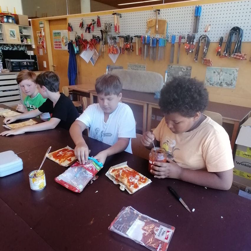Four boys are seated around a table, each preparing a pizza. One boy is adding sauce to his pizza while the others have their toppings ready. Behind them, a wall is filled with various tools and equipment.