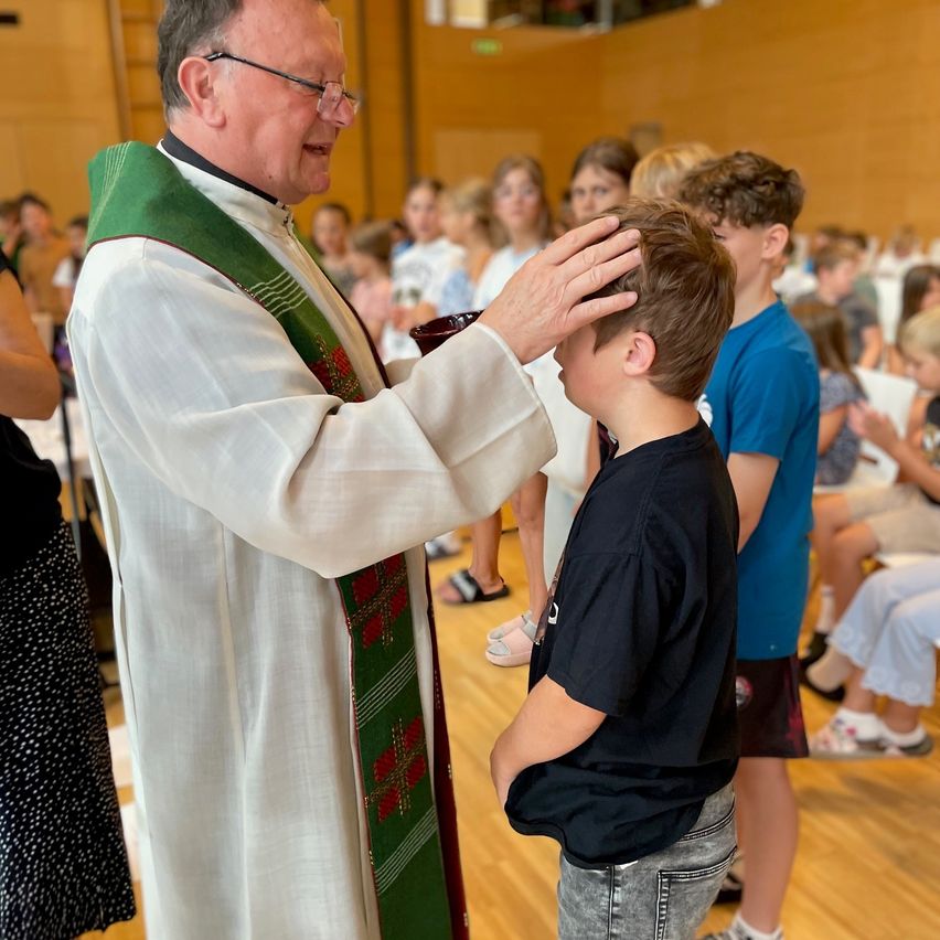A priest in a white robe with a green stole is placing his hand on the head of a young boy during a religious ceremony. The boy looks down, and several other children and adults are seated in the background.
