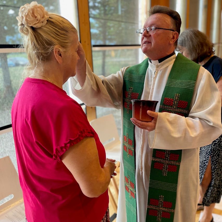 A priest wearing a green and white stole is blessing a woman in a red shirt. They are standing in front of a window with a view of trees outside.