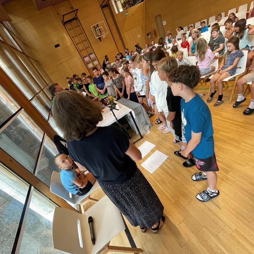 A group of children and adults are gathered in a room. A woman is standing in front of a table with a flower arrangement. The children are standing in line, and some are sitting on chairs. The room has wooden floors and glass windows.