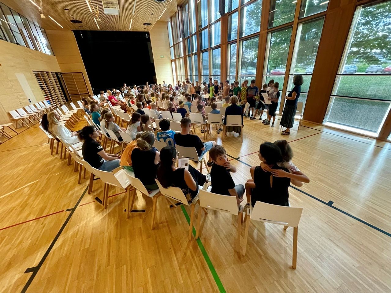 A large group of people, including children, are seated in chairs in a room with large windows. Some are reading papers, and a woman stands at the front.