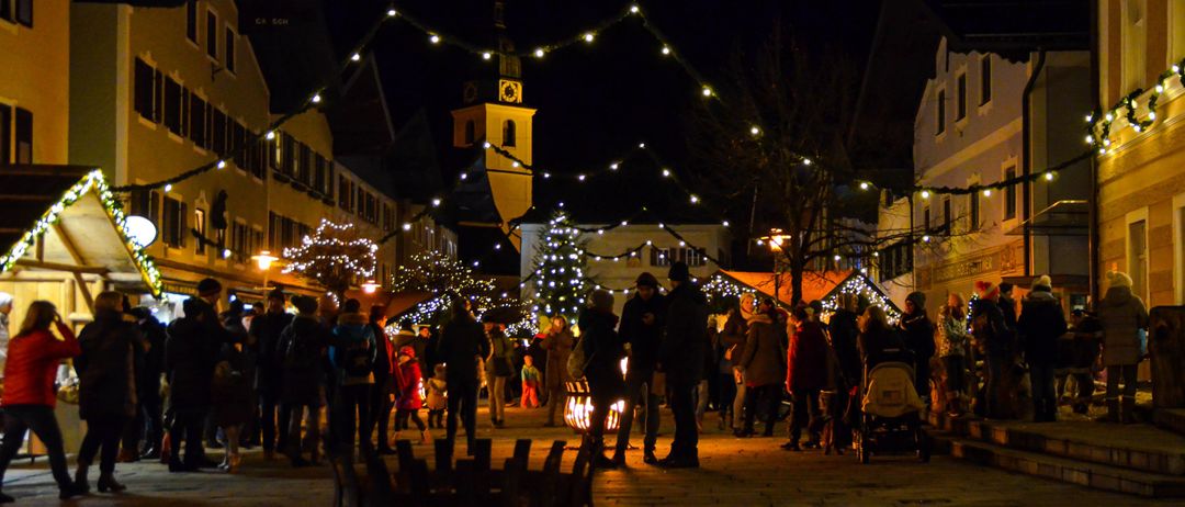 Eine Nachtszene eines festlichen Stadtplatzes mit Menschen, Weihnachtsbäumen und Laternen. Der Platz ist von Gebäuden gesäumt und im Hintergrund befindet sich ein Turm.