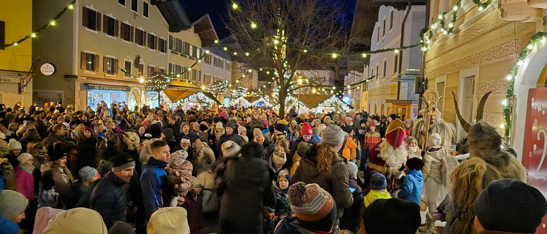 Eine Menschenmenge versammelt sich nachts auf einer Straße, die von Weihnachtslichtern und Ornamenten erhellt wird. Die Straße ist von Gebäuden gesäumt, und in der Mitte steht ein hoher Weihnachtsbaum. Einige Menschen sind in Winterkleidung gekleidet, und ein als Weihnachtsmann verkleideter Mann geht durch die Menge.