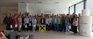 Eine Gruppe von Menschen, die blaue T-Shirts tragen, posiert für ein Foto vor einem Schild, das 'Beach Club' steht. Sie halten weiße Taschen und stehen auf einem gefliesten Boden.