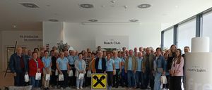 Eine Gruppe von Menschen in blauen Shirts steht in einem Raum mit einem Beach Club-Schild und hält weiße Taschen. Stühle und eine Couch sind in der Nähe.