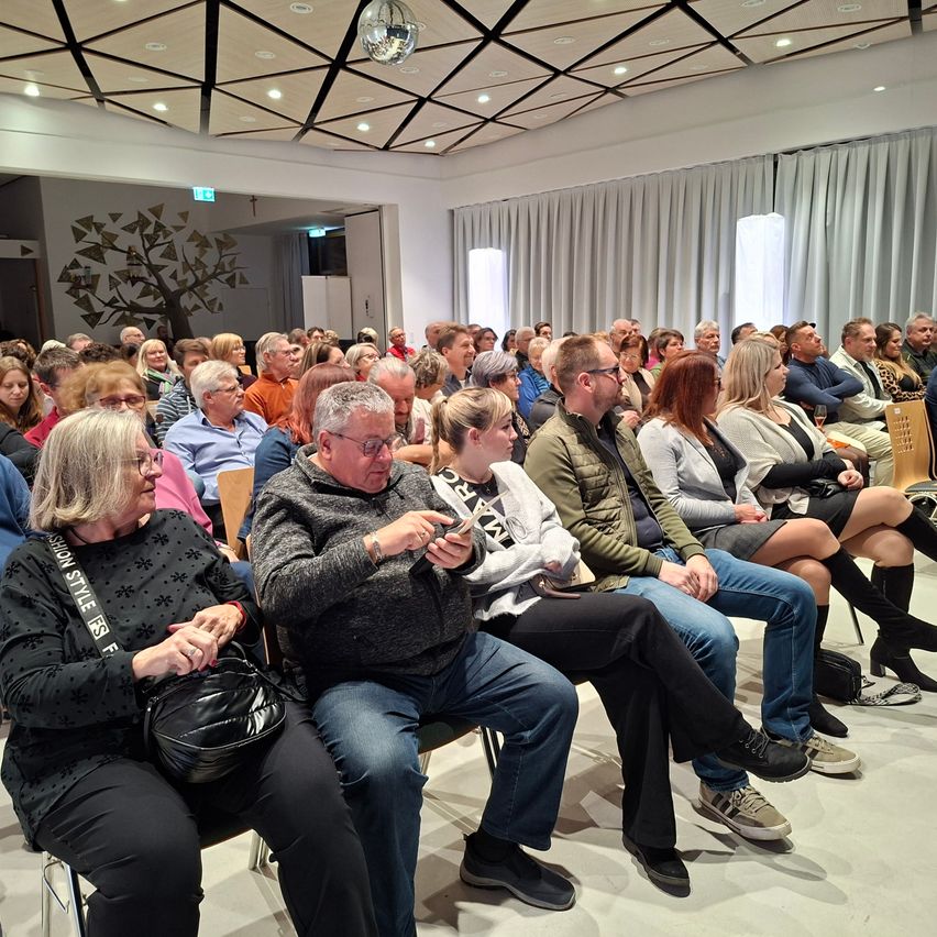 Eine große Gruppe von Menschen sitzt in Stühlen in einem Raum mit einer dreieckigen Decke. Viele tragen Brillen und Turnschuhe. Einige sind mit ihren Handys beschäftigt. An der Wand befindet sich eine Baummalerei.