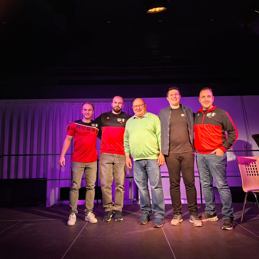 Fünf Männer in passenden Outfits stehen auf einer Bühne unter lila Beleuchtung und lächeln für ein Foto. Sie tragen rote, grüne und schwarze T-Shirts, wobei einer der Männer eine Brille trägt.