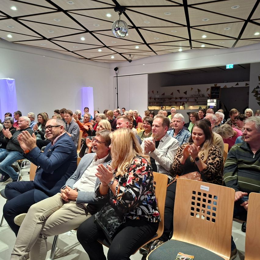 Eine Gruppe von Menschen sitzt in Stühlen in einem Raum, klatscht und lächelt. Der Raum hat ein geometrisches Deckendesign, und eine Discokugel hängt daran. Einige Leute tragen Brillen und Turnschuhe.