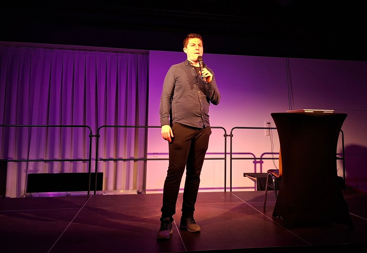 Ein Mann steht auf einer Bühne und hält ein Mikrofon in der Hand. Er trägt ein dunkles Langarmhemd und schwarze Hose. Hinter ihm befindet sich ein violetter Hintergrund mit Vorhängen und einer Metallbarriere.