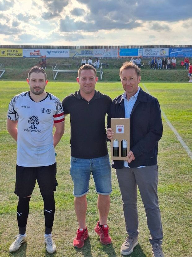 Drei Männer stehen auf einem Rasenfeld mit einer Sporteinrichtung im Hintergrund. Der Mann links trägt ein Trikot und der auf der rechten Seite hält eine Schachtel mit zwei Flaschen.