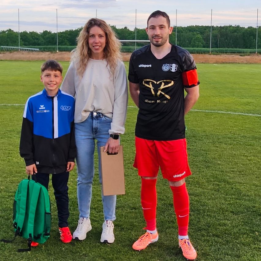 Eine Frau und ein Mann stehen auf einem Fußballfeld mit einem Jungen, der einen grünen Rucksack hält. Der Mann trägt ein schwarzes Trikot mit goldenen Buchstaben und roten Shorts. Der Junge trägt eine blaue und schwarze Jacke mit roten Turnschuhen.